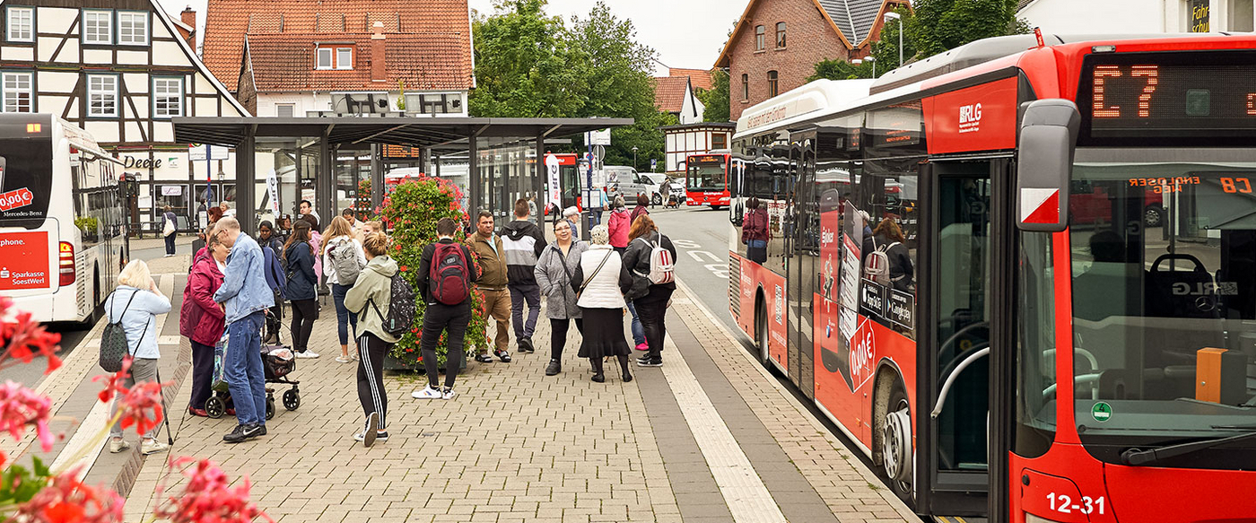 Busbahnsteig mit Menschen. Im Hintergrund die Stadt.