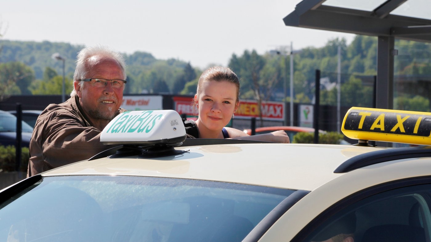 Ein Mann und eine junge Frau stehen hinter einem Taxi. Sie schauen über das Dach des Wagens, auf dem ein Schild TaxiBus zu sehen ist.